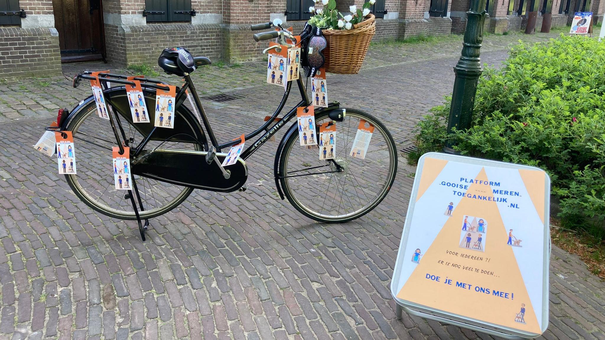 Zwarte damesfiets met rieten mandje. In het mandje witte bloemen. Aan de fiets hangen papieren labels waarop aandacht gevraagd wordt voor het vrijlaten van de openbare ruimte. Voor de fiets een bord met de naam van ons platform en de vraag: Doe je ook mee??